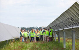 A group of people wearing high visual vests stand between two rows of ground-mounted solar panels.