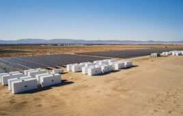 White energy storage units containing electric vehicle batteries sit parallel to a field of solar panels.