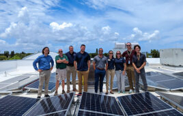 A group of nine people stand shoulder to shoulder between panel rows of a rooftop solar project.