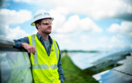A solar installer wearing a hardhat, sunglasses and a high-visual vest leans on an installed row of solar panels.