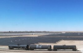 Train cars pass by a field of solar panels on a dry landscape.
