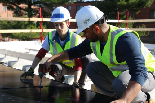Solar installers wearing white helmets and reflective vests use a drill to secure panels to the roof.