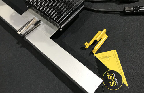 A yellow clip with jagged edges, a business card and a module-level power electronic unit attached to a solar panel frame rest upon a black tablecloth. 