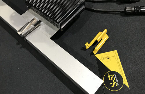 A yellow clip with jagged edges, a business card and a module-level power electronic unit attached to a solar panel frame rest upon a black tablecloth.